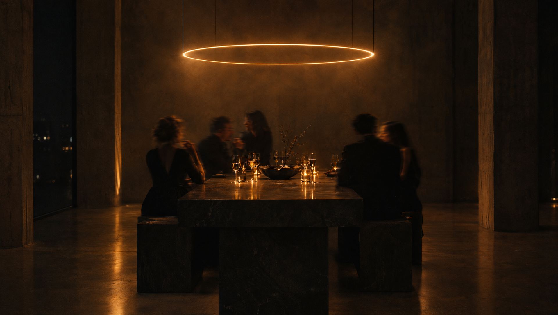 Des convives autour d'une table de pierre, sous un anneau de lumière, dans un espace de béton